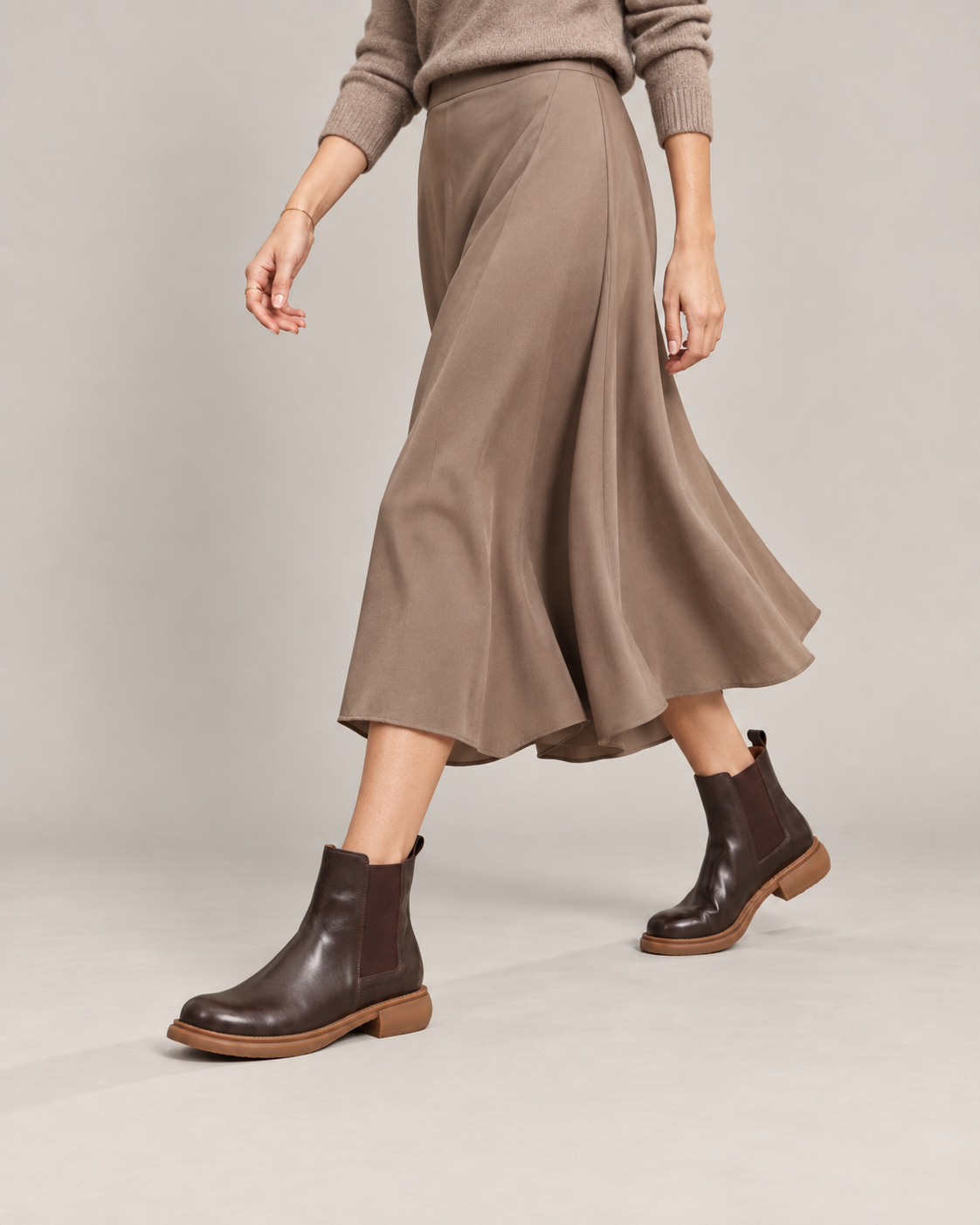 Brown leather Chelsea boots worn in a walking pose with a flowing taupe midi skirt and matching sweater.