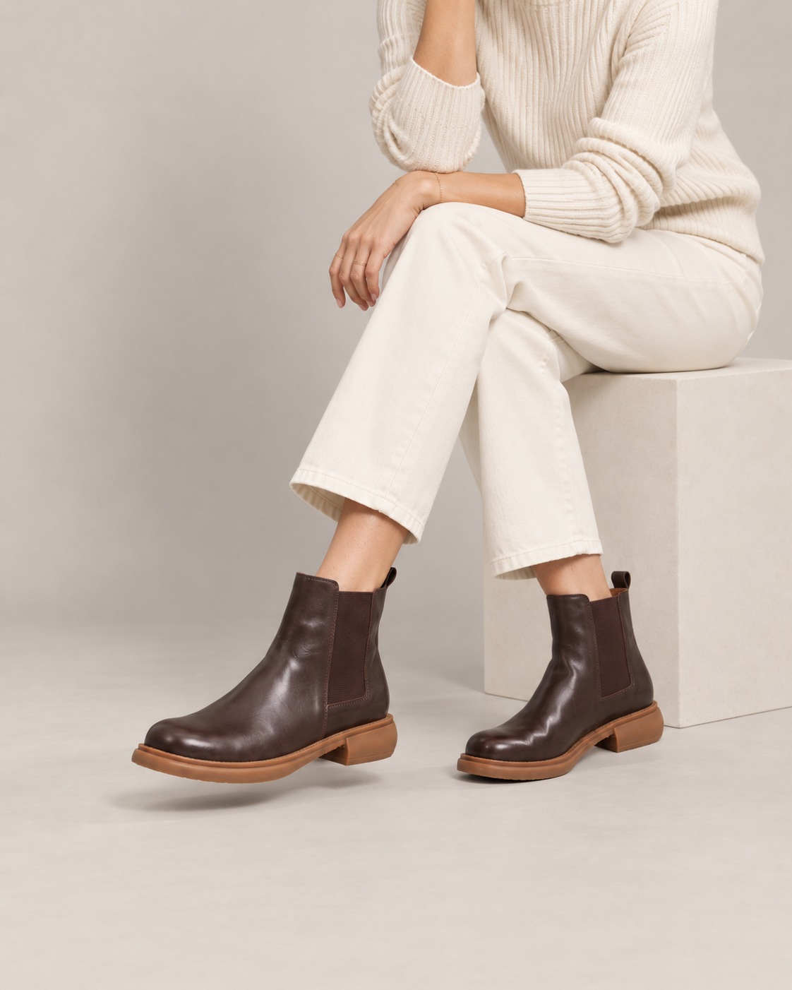 Close on-foot view of brown leather Chelsea boots worn with cream trousers by a seated model.