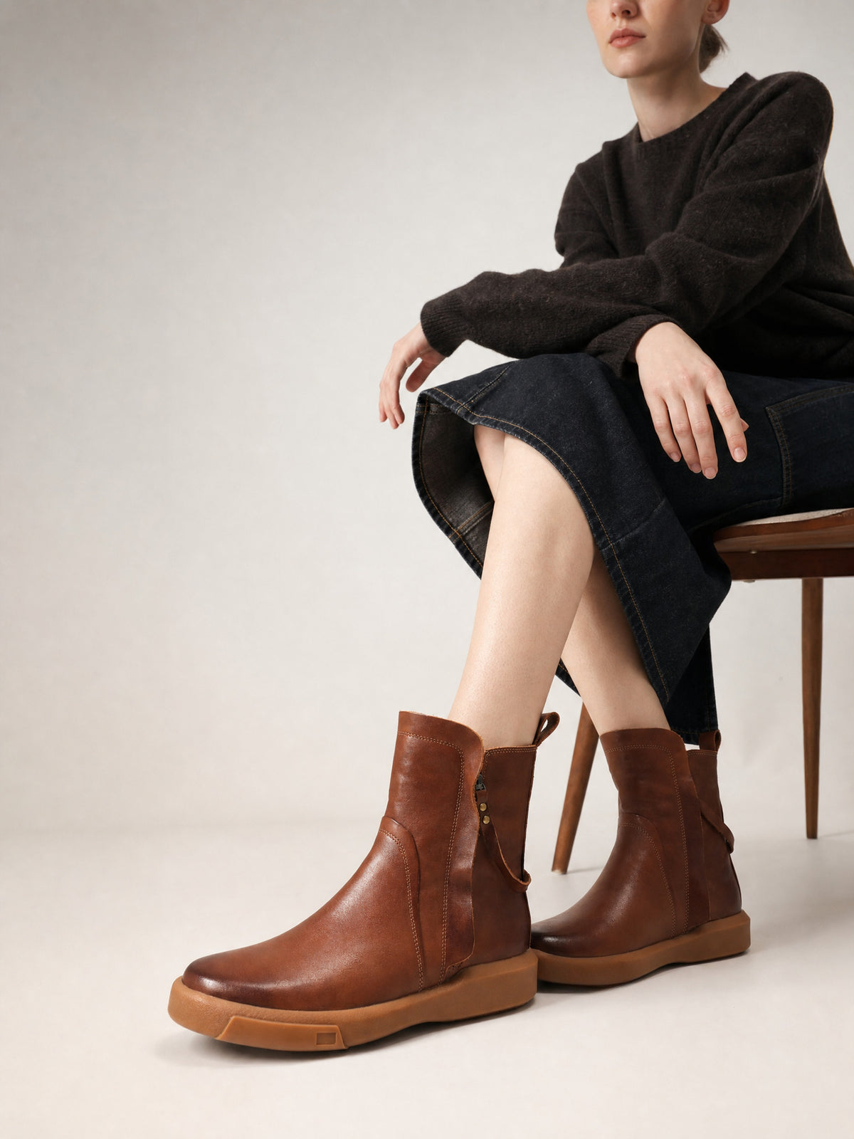 Seated model wearing brown leather ankle boots with a black sweater and dark denim midi skirt.